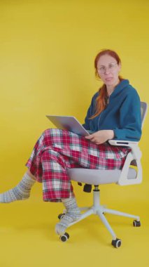 Pijamalı ve gözlüklü bir kadın evden bilgisayarda çalışıyor. Ofis sandalyesinde oturuyor.