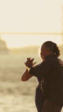 Nehir köprüsünün yanında dans eden yaşlı bir çift. Arka planda büyük bir yolcu gemisi, gün doğumu.