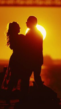 Gün batımının yumuşak renkleri ve arkaplanda köprü olan bir çift arasında samimi bir kucaklaşma yakalar..