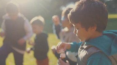 Parkta güneşli bir günde dışarıda sabun köpüğüyle oynayan bir grup mutlu çocuk.
