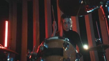 Genç bir kadın, karanlık bir stüdyoda kırmızı ve mavi sahne ışıklarıyla enerjik bir şekilde davul çalıyor.