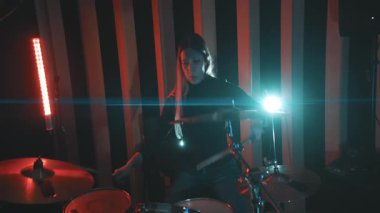Dramatik kırmızı ve mavi ışıklandırmalı bir stüdyoda davul setinde pratik yapan enerjik bir kadın.