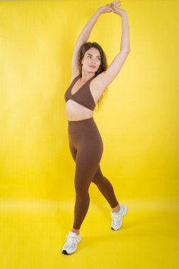 Genç bir kadın esniyor, atletik kahverengi bluz ve tayt giyiyor, sarı arka planda.