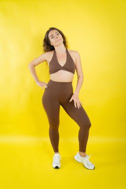 Kahverengi spor sutyen ve tayt giyen kadın model, sarı arka planda güvenle duruyor.