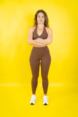 Spor sutyenli ve taytlı bir kadın kollarını kavuşturup doğrudan kameraya bakıyor.