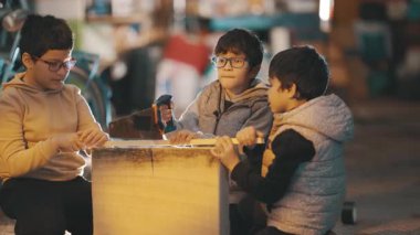 Üç çocuk bir atölyede çalışıyor, bir okul projesi için odun kesiyorlar.