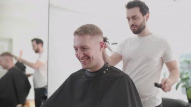 Profesyonel berber, modern bir kuaförde gülümseyen bir müşterinin saçını elektrikli makasla buduyor.