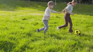 Aktif erkek ve kız kardeşler yeşil çimlerde koşup futbol topunu tekmeliyorlar.