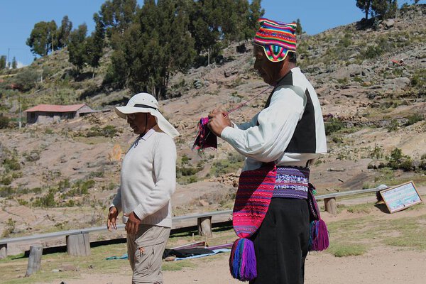 Isla Taquile, Puno, Peru - May 29, 2025: Male Taquileo presents his knitting workpiece hanging around his neck. Colorful clothes with chullo and coca leaves pouch on belt. Tourguide translates.