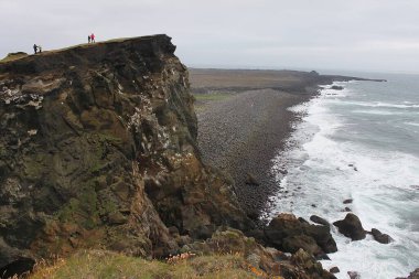 Valahnukamol sahilindeki dramatik sarp kayalıklar Atlantik Okyanusu 'nun dalgalarını ve Reykjanes yarımadasındaki gri gökyüzünün altındaki siyah çakıl taşı plajlarını seyreden turistlerle dolu. Doğanın gücü.