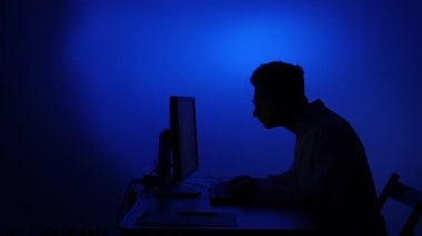 Masada oturan siluetli bir adam, yoğun bir şekilde bilgisayar ekranına odaklanmış, gece siber güvenliği, kodlamayı ya da uzaktan çalışmayı temsil eden mavi bir ışık tarafından aydınlatılmış.