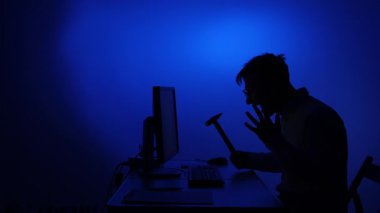 Silüet içindeki bir adam, bir bilgisayar monitörüne çekiçle vurmaya hazırlanırken hayal kırıklığını ve kızgınlığını ifade ediyor. Koyu mavi arka planda stresi ve teknolojik zorlukları simgeliyor.