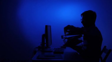 Koyu renk siluet, elinde bir bilgisayarla bir masada otururken uzun bir yığın klasör taşıyor. Mavi ışıklı ofis ortamında iş stresi ve bürokratik aşırı yüklemeyi temsil ediyor.