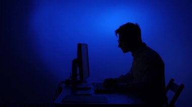 Mans silueti hayal kırıklığı içinde bir bilgisayar klavyesine vuruyor, dramatik mavi ışıklı arka plana karşı işyeri stresi, öfke ve teknik sorunları sembolize ediyor