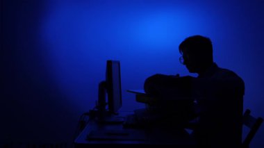 Silüette çalışan ya da bilgisayar monitörüyle çalışan, canlı mavi ışıkla aydınlatılan, teknolojiyi, konsantrasyonu ya da gizemli online aktiviteyi sembolize eden kişi.