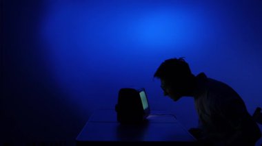 Siluetteki adam karanlık bir odada parlayan bir retro monitöre doğru eğiliyor, klasik crt ekrandan mavi ışıkla yıkanmış, odaklanmış ve iç gözlem altında yalnız.