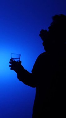 Dikey videocuların koyu renk silueti, parmakları ve bir bardak su arasında küçük bir hap tutuyor, mavi arka planda ilaç almaya hazırlanıyor, sağlık ve tedaviyi simgeliyor.