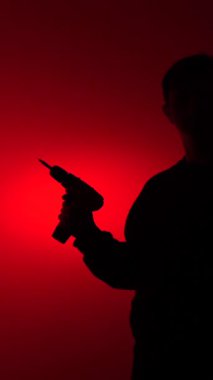 vertical video Man silhouette holding a power drill, creating a menacing and dangerous mood under a bright red spotlight, emphasizing anonymous threat and hidden intent