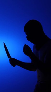 Dikey video kapüşonlu bir adam keskin bir bıçak tutuyor dramatik mavi ışık altında, tehlike, korku ve şiddeti sembolize ediyor.