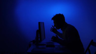 Mans siluet çığlık ve bir bilgisayar ekranı elleri ile işaret, yoğun hayal kırıklığı, stres ve öfke ifade mavi ışık ile karanlık bir odada
