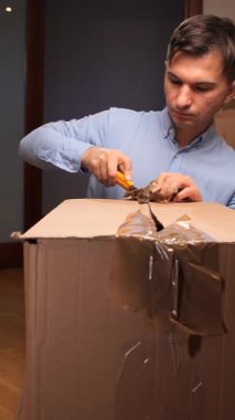 Dikey video Genç adam kapalı bir karton teslimat kutusunu bıçak ile açıyor, taşındıktan ya da internetten sipariş aldıktan sonra içindekileri açıyor, ellerini ve kutusunu kapatıyor.