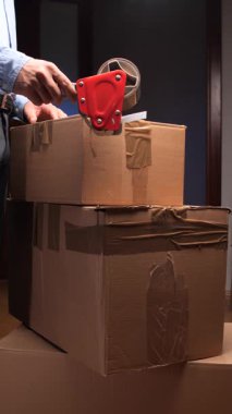 dikey video Kişi mavi gömlek ve gri pantolon giyerek bir karton kutuyu mühürlemek için kırmızı bant makinesi kullanır, onu bir ev veya ofis ortamında nakliyeye ya da taşınmaya hazırlar.