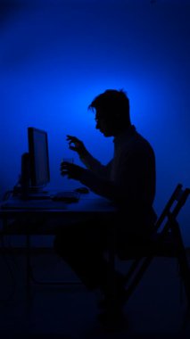 Gece geç saatlerde elinde bir hap ve bir bardak su tutan, uykusuzluk, anksiyete, ilaç kullanımı ve izolasyon taşıyan, bilgisayar masasındaki bir adamın dikey video silueti.