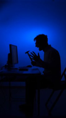 Bir bilgisayar monitörüne çekiçle vurmaya hazırlanırken hayal kırıklığını ve öfkeyi ifade eden silüet içindeki dikey videocu adam, koyu mavi arka plana karşı stresi ve teknolojik zorlukları sembolize ediyor.