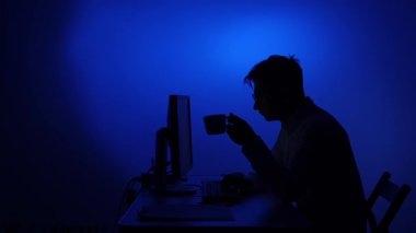 Masaüstü bilgisayarında çalışan, koyu mavi ışıklı bir odada kahve içen, gece geç saatlere kadar çalışma, uzaktan kumandalı işler ve siber güvenlik kavramlarını temsil eden silüetli bir adam.