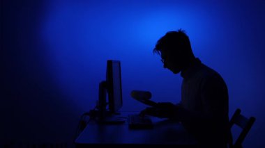 Masadaki koyu renk siluet bir adam, bir panoya yazmaya odaklanırken mavi ışıklı bir bilgisayar ekranı karanlık bir ev ofisinde parlıyor, uzaktan kumandalı iş sahnesinde.