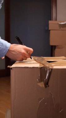 Taşınmak için paketleme yaparken veya paket hazırlarken karton bir kutuya kargo adresi yazan dikey video adam, kapalı bir ayarda elleri ve etiketleri kapatın