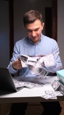 Dikey video. Mutlu genç adam bir masada oturuyor ve mali başarıyı kutluyor, elinde büyük miktarda para tutuyor, karı, geliri, kazancı ve tasarrufu temsil ediyor.