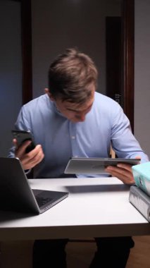 Dikey video stresli erkek çalışan ağır iş yükünden bunalmış, akıllı telefon ve tableti bir masanın üzerinde dizüstü bilgisayar ve klasörler ile iş yerinin tükendiğini gösteren bir yığın resimle tutuyor.