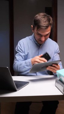 dikey video genç adam akıllı telefon kullanarak iletişim kurarken gülümsüyor, dijital tablet tutuyor, dosyalarla dolu bir masada dizüstü bilgisayar üzerinde çalışıyor, yoğun uzaktan çalışmayı ve modern iletişimi temsil ediyor. İnsan teknolojiyi kullanarak aynı anda birden fazla iş yapar. 