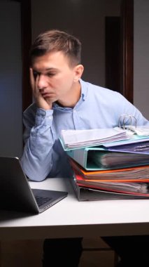 Dikey video Genç adam hayal kırıklığıyla başını tutuyor, bir dizüstü bilgisayarla bir masada oturuyor ve bir yığın renkli dosya ve evrak, ağır bir iş yükü ve tükenmişliği temsil ediyor.