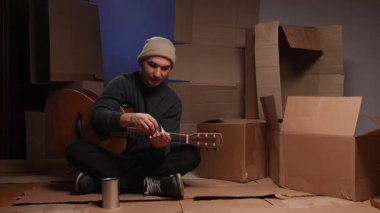 Evsiz bir adam akustik gitar çalıyor ve gülümsüyor. Etrafı boş kutularla çevrili karton çarşafların üzerinde otururken para alıyor. Yoksulluğu ve bağışları temsil ediyor.