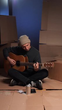 Dikey video, evsiz adam karton kutularda oturuyor, akustik gitar çalıyor, bağış yapmak için bir bardak ve akıllı telefon, yoksulluğu, sokak sanatını ve dayanıklılığı simgeliyor.