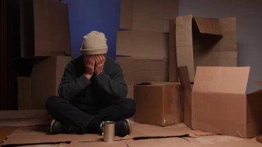 Evsiz bir adam karton kutularda oturuyor, elleriyle yüzünü kapatıyor, sokakta para dilenirken üzüntü, yoksulluk ve umutsuzluk duygularını ifade ediyor.