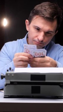 Dikey Videocu, eski bir daktilo ve boş bir kağıtla bir masada otururken, bir yazarlık mesleğinin mali başarısını ve ödülünü simgeleyen yığınla dolarımızı sayıyor.