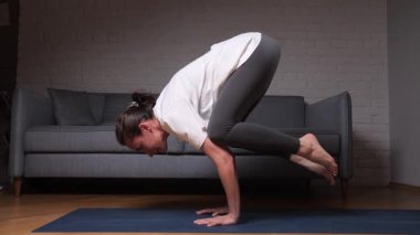 Bakasana yogasında dengede duran bir kadın, mavi bir minder üzerinde duruyor, bir ev antrenmanı sırasında güç, odaklanma ve farkındalık için kol dengesi egzersizi yapıyor.