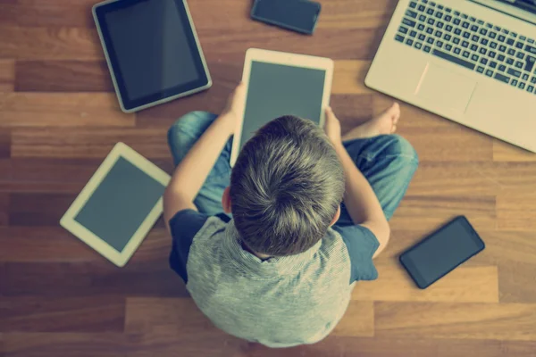 Little boy using tablet sitting on wooden floor. Digital tablets, laptop, mobile phones all around. Selective focus. Classic toned image. Top view. Education, learning, technology concept