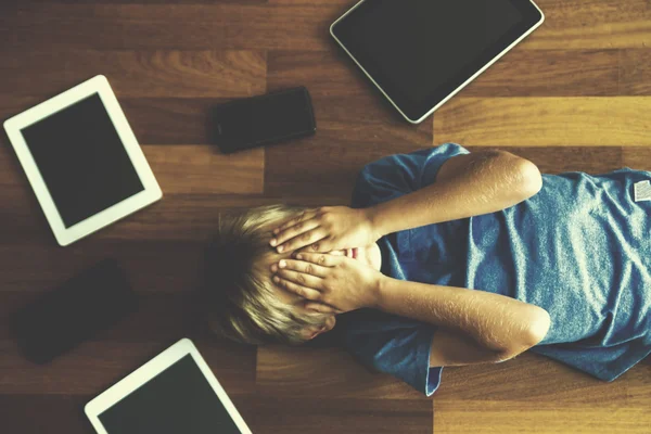 Bambino tenendo tablet sopra la testa sdraiato sul pavimento in legno. Tablet digitali, telefoni cellulari, laptop ovunque. Vista dall'alto. Istruzione, apprendimento, concetto tecnologico. Immagine tonica . — Foto Stock