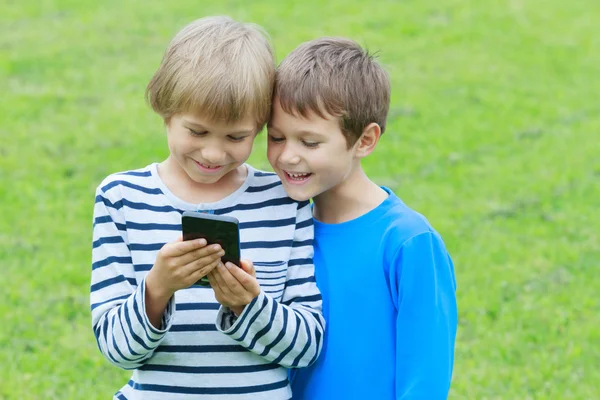 A gyermekek mobiltelefon kültéri. Két fiúk, mosolyogva, látszó-hoz képernyő, játék vagy alkalmazás segítségével. Technológia oktatás szabadidős emberek koncepció — Stock Fotó