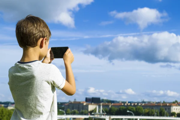 Niño con teléfono móvil. Niño tomando fotos de su teléfono inteligente. Hermoso cielo y fondo de la ciudad. Vista trasera. Concepto tecnológico — Foto de Stock
