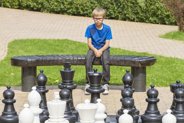 Bambino concentrato, pensando alla sua prossima mossa, seduto su una panchina di legno durante una partita a scacchi all'aperto usando pezzi a grandezza naturale e tavola — Foto Stock