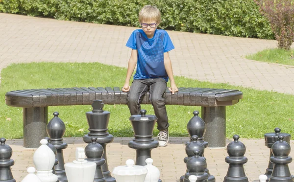 Niño concentrado, pensando en su próximo movimiento, sentado en un banco de madera durante un juego de ajedrez al aire libre usando piezas de tamaño natural y tablero —  Fotos de Stock