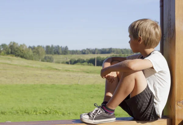 Niño sentado en la terraza al aire libre y sueña o piensa en algo. Día de verano. Fondo de prados verdes — Foto de Stock