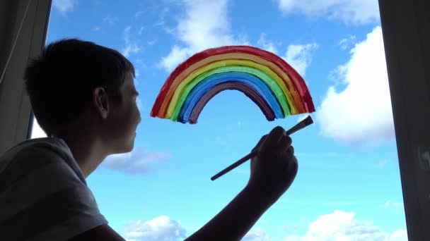 Мальчик рисует разноцветную радугу на окне во время карантина Ковид-19 дома. Оставайтесь дома из-за опасности коронавирусной инфекции. Символ надежды. 4k — стоковое видео