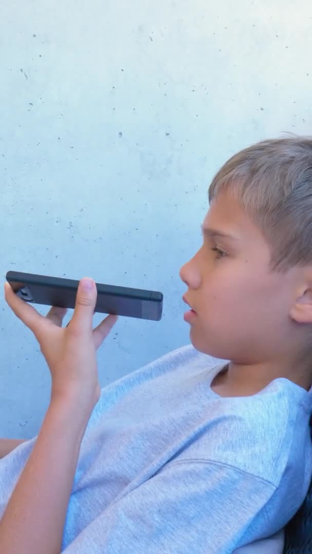 Το αγόρι χρησιμοποιεί αναγνώριση φωνής στο έξυπνο τηλέφωνο. Βίντεο με κάθετο προσανατολισμό οθόνης. Βίντεο 4k — Αρχείο Βίντεο
