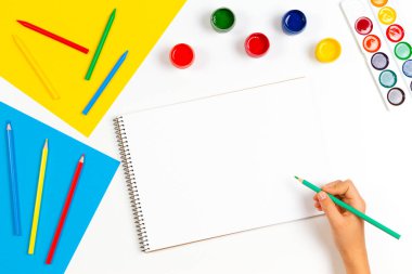 Çizim defterinin üzerinde renkli kalemler ve resim malzemeleriyle çocuk eli çizimi. Öğrenme, hobi, sanat geçmişi. Üst görünüm